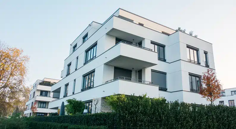 Straßenseitige Ecke eines weißen modernen Mehrfamilienhauses mit Balkonen Flachdach und Heckenbegrünung