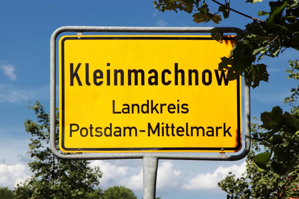 Frontal aufgenommener gelber Ortseingangsschild Kleinmachnow mit schwarzer Umrandung vor blauem Himmel und Bäumen