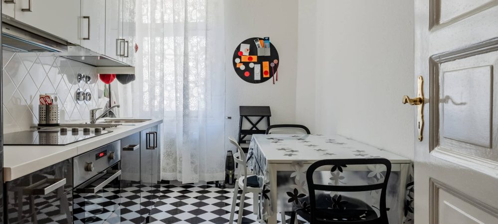 Innenansicht kompakte Küche in Wohnung aus Türperspektive mit schwarzweißen Bodenfliesen, Tisch, Fenster mit Gardine