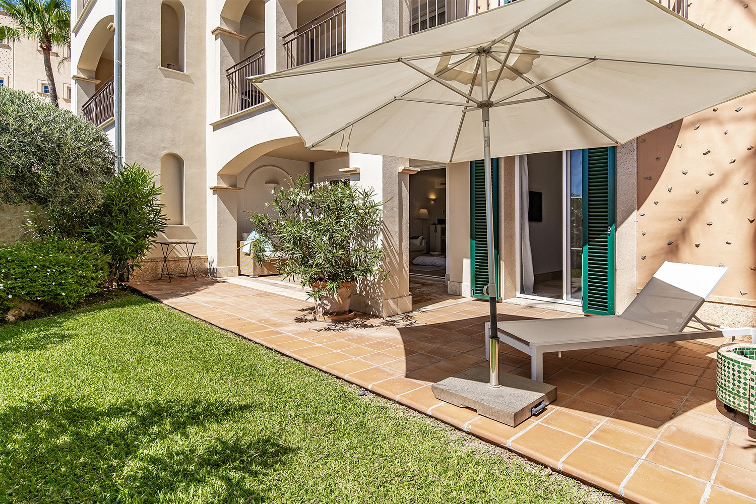 Blick von Garten auf Erdgeschosswohnung mit Terrasse, Sonnenschirm, Liegeliege und mediterraner Fassade mit Holzläden