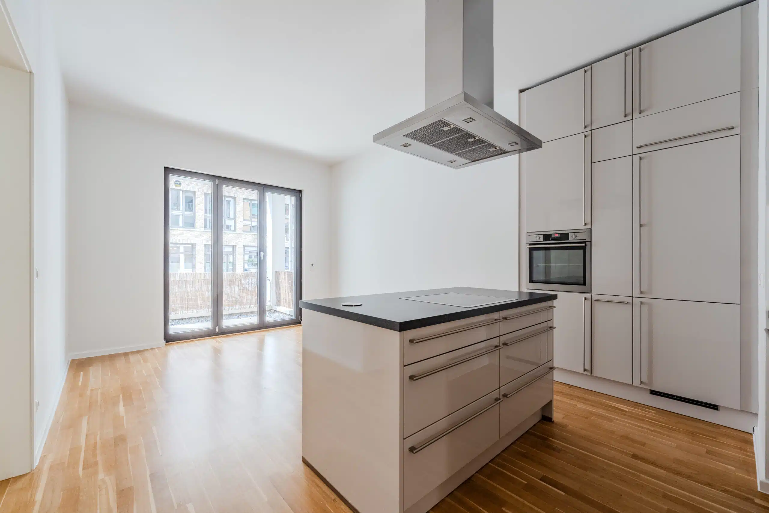 Innenansicht offene Küche in Neubauwohnung mit Kücheninsel, Dunstabzugshaube, Parkettboden und bodentiefen Fenstertüren